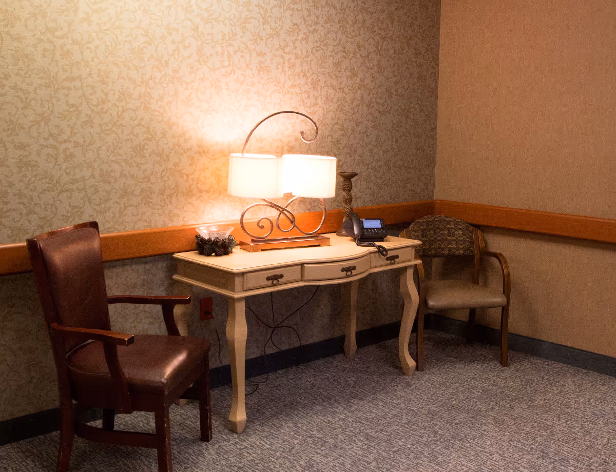 Small seating nook with a decorative table holding a lamp and phone, flanked by two chairs against patterned wallpaper.