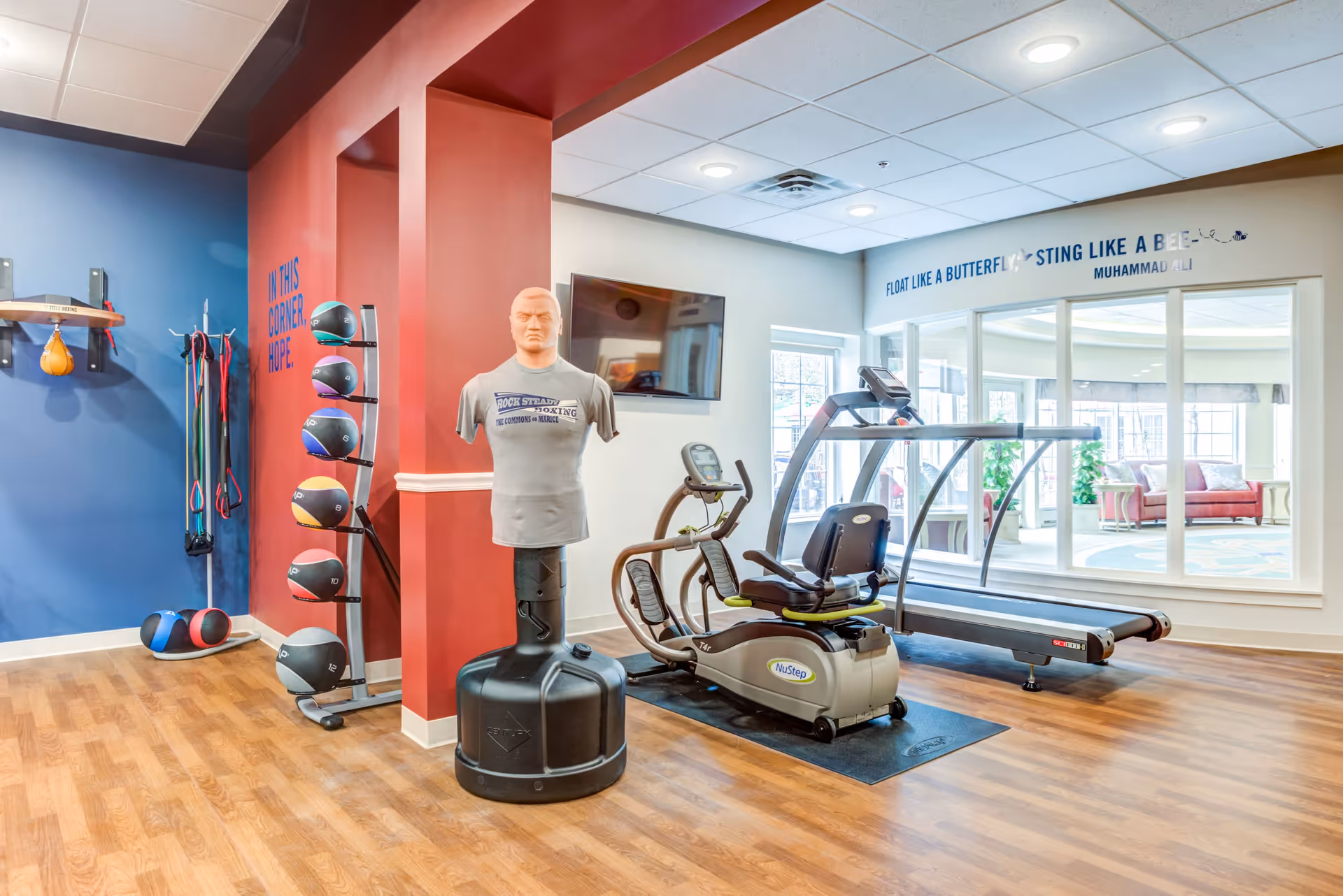 A fitness room with wooden flooring featuring a punching bag mannequin, a treadmill, a recumbent exercise bike, and a rack of medicine balls. The walls are painted blue and red, with a motivational quote on the wall that reads 'FLOAT LIKE A BUTTERFLY, STING LIKE A BEE - MUHAMMAD ALI'. A large window looks into a bright lounge area with sofas and plants.