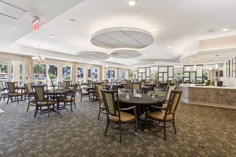Spacious dining room with multiple round tables covered with black tablecloths, each surrounded by wooden chairs with cushioned seats and backs. The room has large windows allowing natural light to fill the space, beige curtains, and modern circular ceiling light fixtures. A counter area is visible on the right side of the room.