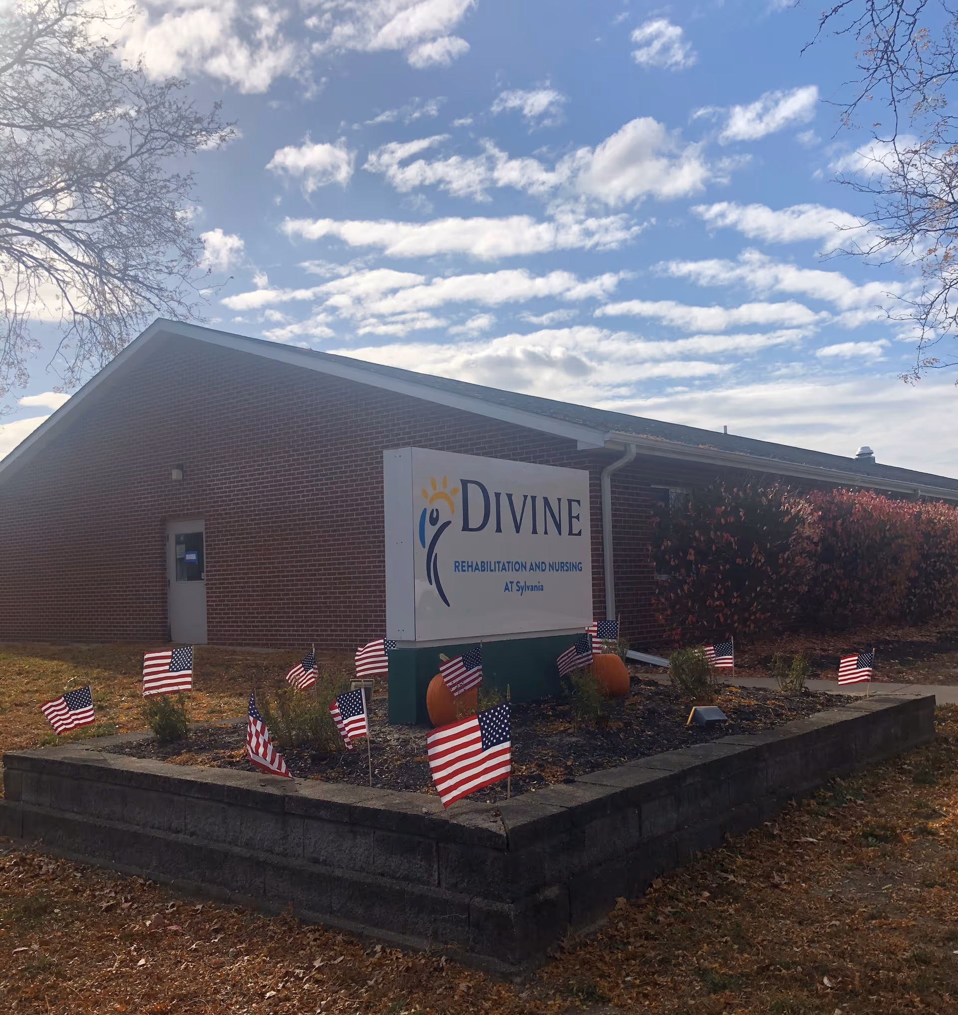 Brick nursing facility exterior with a large 'Divine Rehabilitation and Nursing at Sylvania' sign surrounded by small American flags and pumpkins.