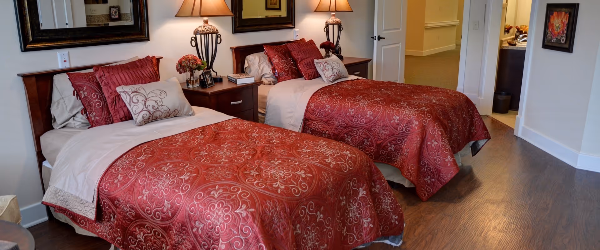 A senior living facility bedroom with two twin beds, each adorned with red patterned bedspreads and multiple pillows. Between the beds is a wooden nightstand with a decorative lamp, flowers, and a book. The room has wooden flooring, a mirror above the nightstand, and an open door leading to a bathroom area.