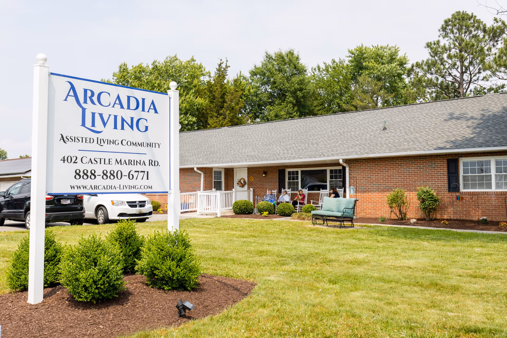 Front lawn and large sign for Arcadia Living assisted living community in front of a single-story brick building with benches and parked cars.