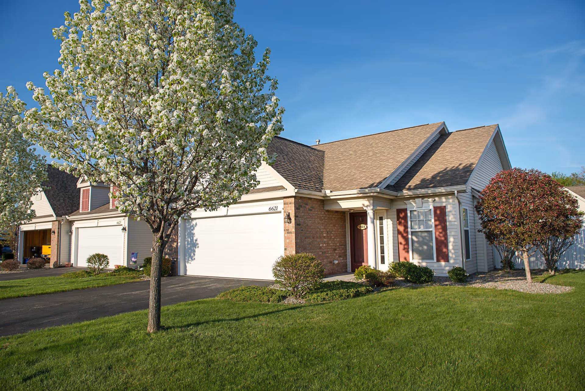 Exterior view of a single-story residential building with a two-car garage, a front door with sidelights, and windows with red shutters. The house is surrounded by a well-maintained green lawn, flowering trees, and shrubs under a clear blue sky.