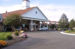 Front entrance of a retirement center with a covered porte-cochere, driveway, parked car, and landscaped grounds.