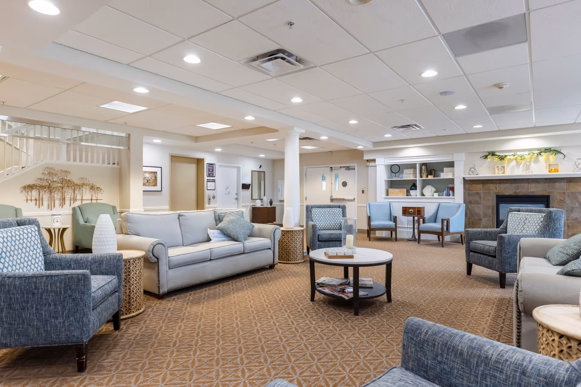 A spacious and well-lit living room area in a senior living facility with multiple comfortable armchairs and sofas arranged around a central round coffee table. The room features a carpeted floor, a fireplace with decorative items on the mantel, built-in shelves with decor, and soft lighting from recessed ceiling lights. There is a staircase visible in the background and a few side tables with lamps and decorative objects.