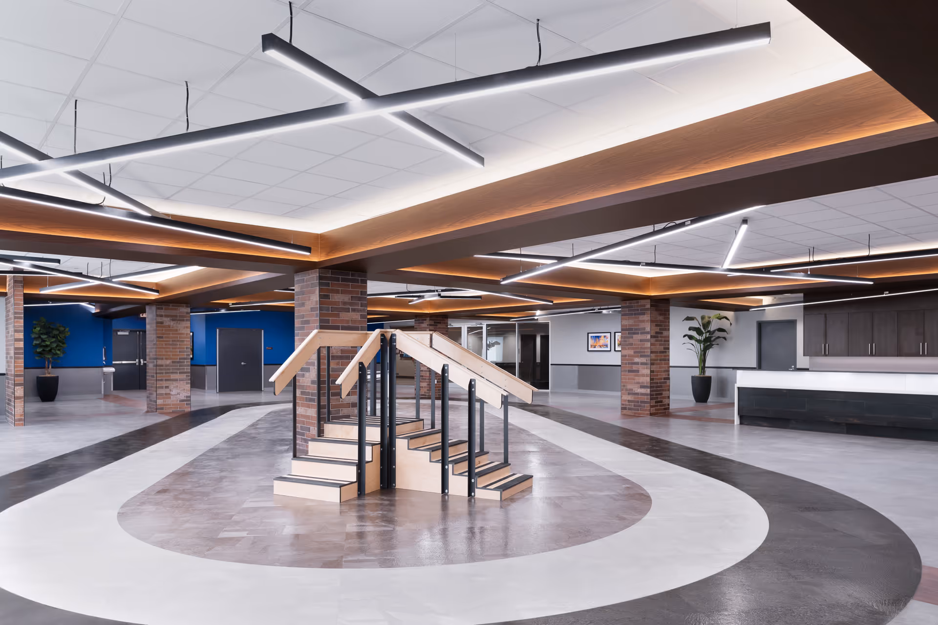 A spacious, modern interior of a rehabilitation or nursing facility with a central wooden staircase designed for physical therapy. The room features brick columns, blue accent walls, contemporary linear ceiling lights, and a reception desk with dark cabinetry. There are plants and framed artwork on the walls, creating a welcoming atmosphere.