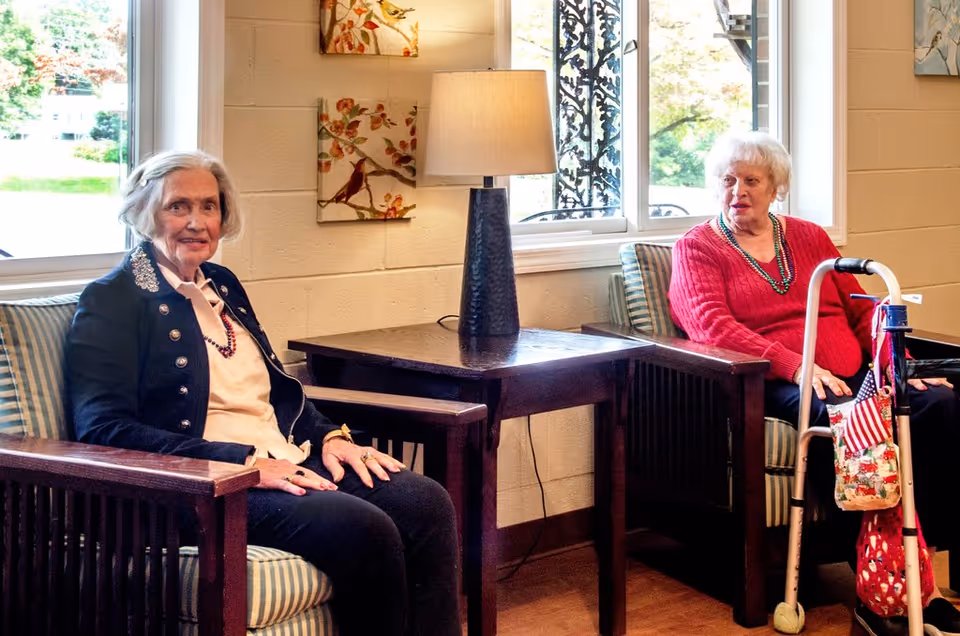 Two elderly women sitting in a cozy living room area with wooden armchairs and striped cushions. A table with a lamp is placed between them, and there are windows behind them letting in natural light. One woman is wearing a black jacket and the other a red sweater, with a walker beside her.