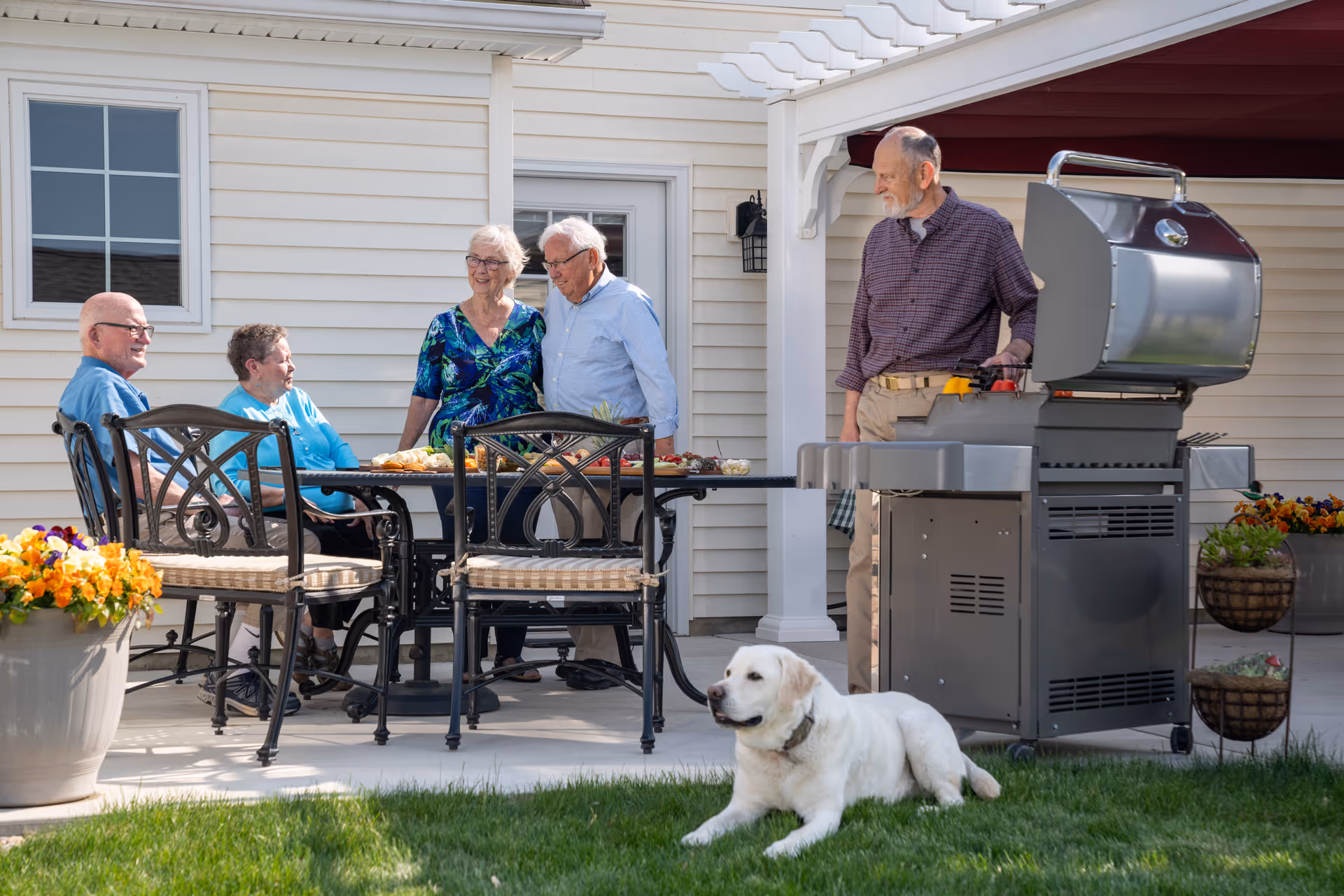 A group of four elderly people socializing around a patio table with food, while a man grills on a barbecue. A white dog lies on the grass nearby. The setting is an outdoor patio area with flower pots and a beige house exterior in the background.