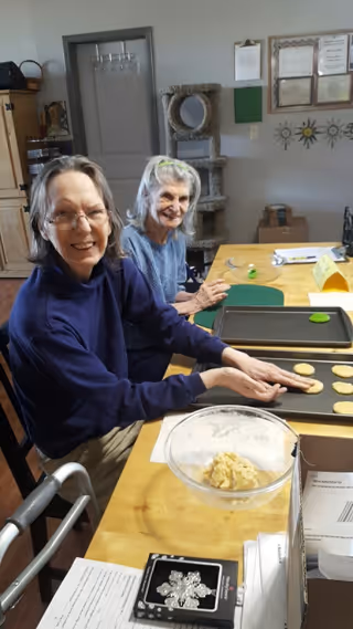 Two elderly women sitting at a table in a room, smiling and placing cookie dough on baking trays. There is a bowl with cookie dough on the table, along with some papers and a walker nearby. The background shows a cat tree, a door, and some framed certificates on the wall.