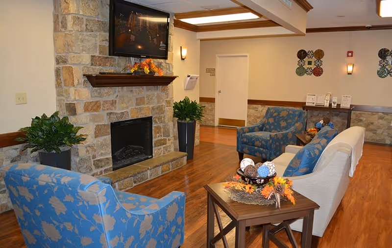 A cozy common room with patterned armchairs, a sofa, a stone fireplace with a mounted TV, and decorative plants and tables.