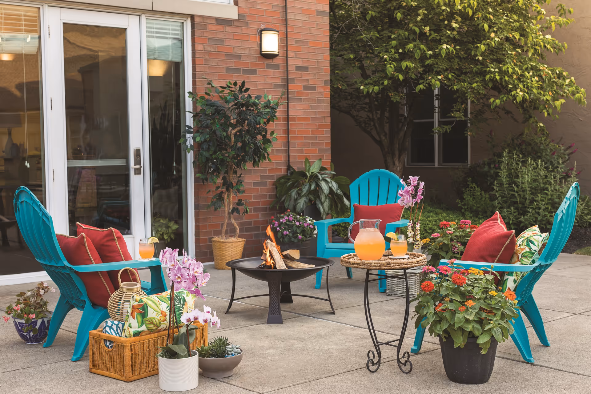 Outdoor patio area with four turquoise chairs arranged around a small fire pit. Each chair has red cushions, and there are various potted plants and flowers placed around the seating area. A small round table holds a pitcher and glasses of orange drink. The patio is adjacent to a brick building with large glass doors and windows.