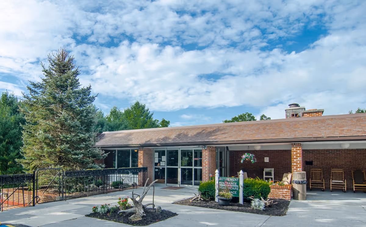 Exterior view of Glenwood Park facility showing a single-story brick building with a covered entrance. There is a sign indicating retirement and healthcare entrances, surrounded by small landscaped areas with plants and flowers. A large evergreen tree is visible to the left, and the sky is partly cloudy.
