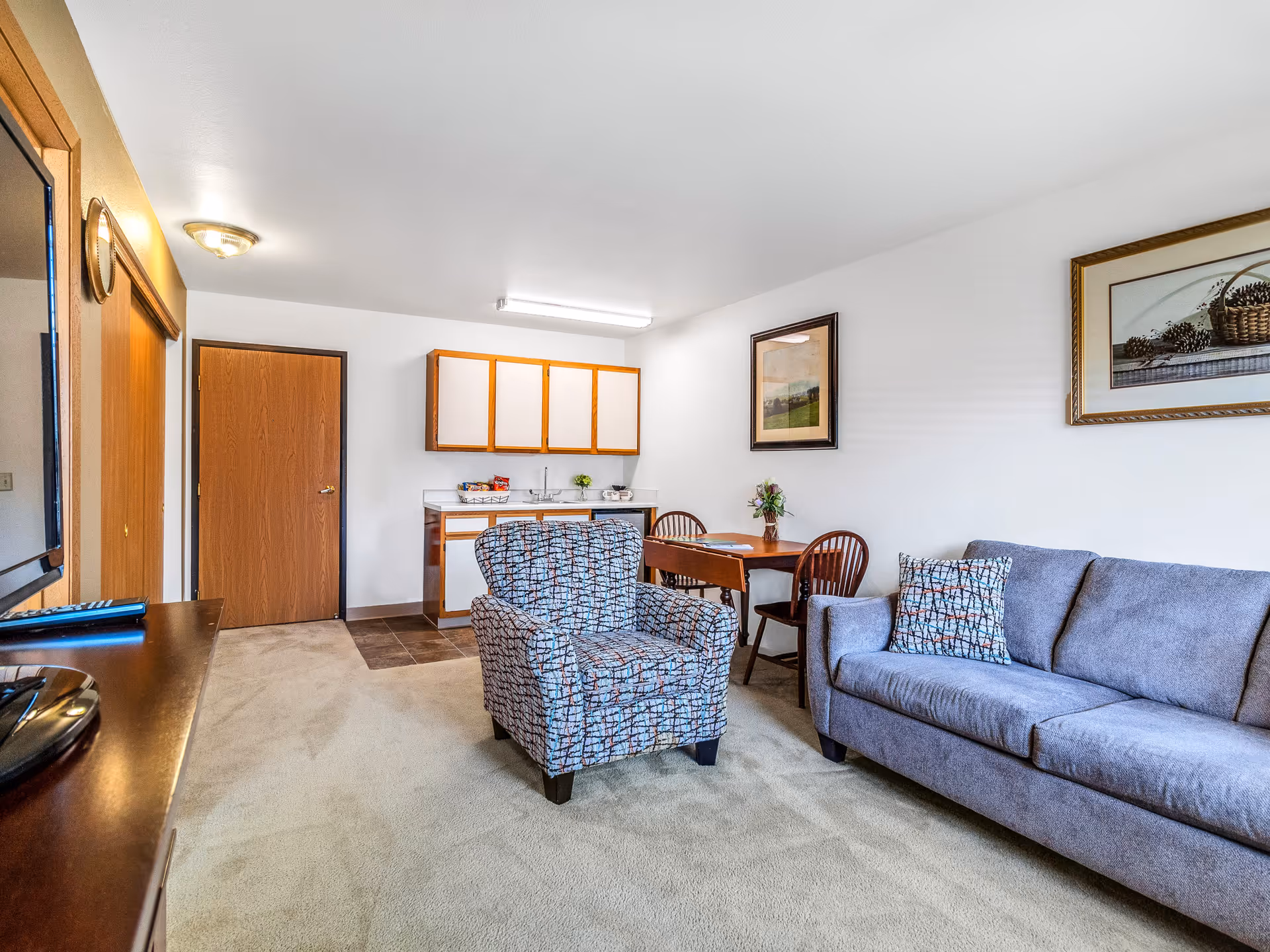 Furnished living area with a gray sofa, patterned armchair, small dining table and a kitchenette in the background.