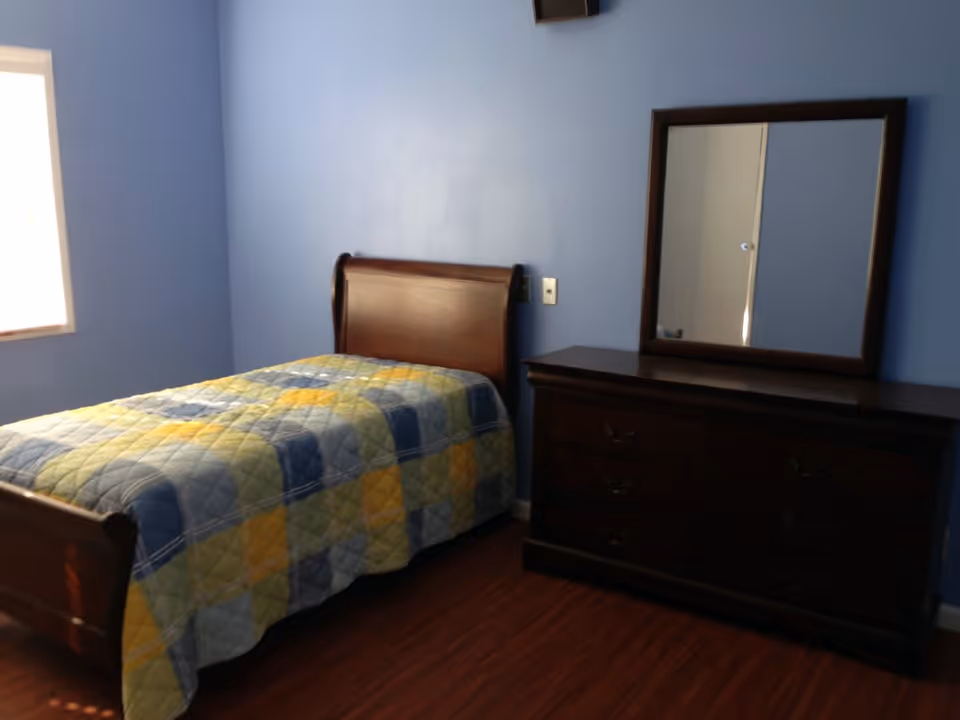 A simple bedroom with blue walls, a wooden bed with a colorful patchwork quilt, and a dark wooden dresser with a mirror on top. The room has a window letting in natural light and wooden flooring.