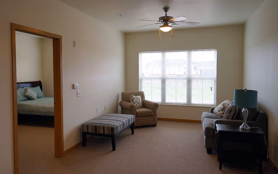 A cozy living room area with a large window letting in natural light. The room features a striped ottoman, a patterned armchair with a decorative pillow, a gray sofa with pillows, and a side table with a blue lamp. A ceiling fan with a light is mounted on the ceiling. To the left, an open doorway leads to a bedroom with a bed and pillows visible.