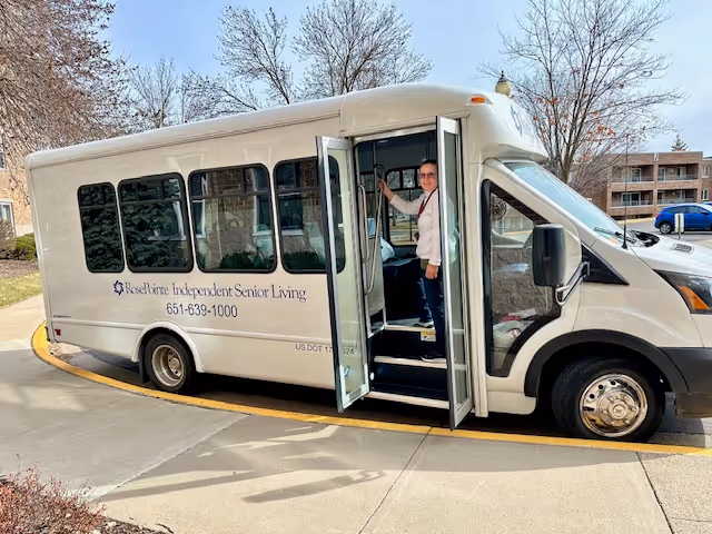 A white shuttle bus with the text 'RosePointe Independent Senior Living 651-639-1000' on the side is parked on a curved driveway. The bus door is open, and a person is standing inside near the entrance. Trees and a building are visible in the background under a clear sky.