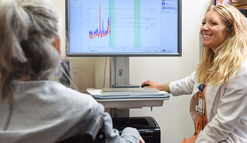 A healthcare professional with a name badge and white coat is smiling and pointing at a computer screen displaying a graph, while interacting with a person in a wheelchair in a clinical setting.