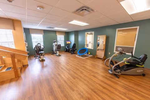 A spacious rehabilitation room with wooden flooring and green walls, equipped with exercise machines including stationary bikes and a recumbent bike. There is a small trampoline, a mirror, and a wooden staircase with handrails on the left side. Large windows allow natural light to enter the room.