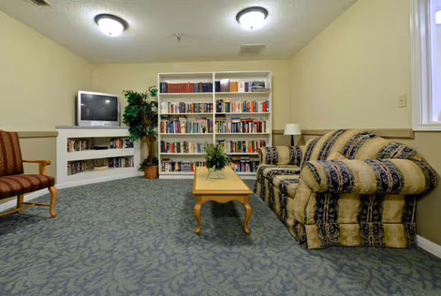 A cozy living room area with a patterned sofa and a wooden coffee table with a plant centerpiece. There is a bookshelf filled with books against the wall, a small TV on a corner shelf, a potted plant, and a single upholstered chair. The room has carpeted flooring and beige walls with two ceiling lights.