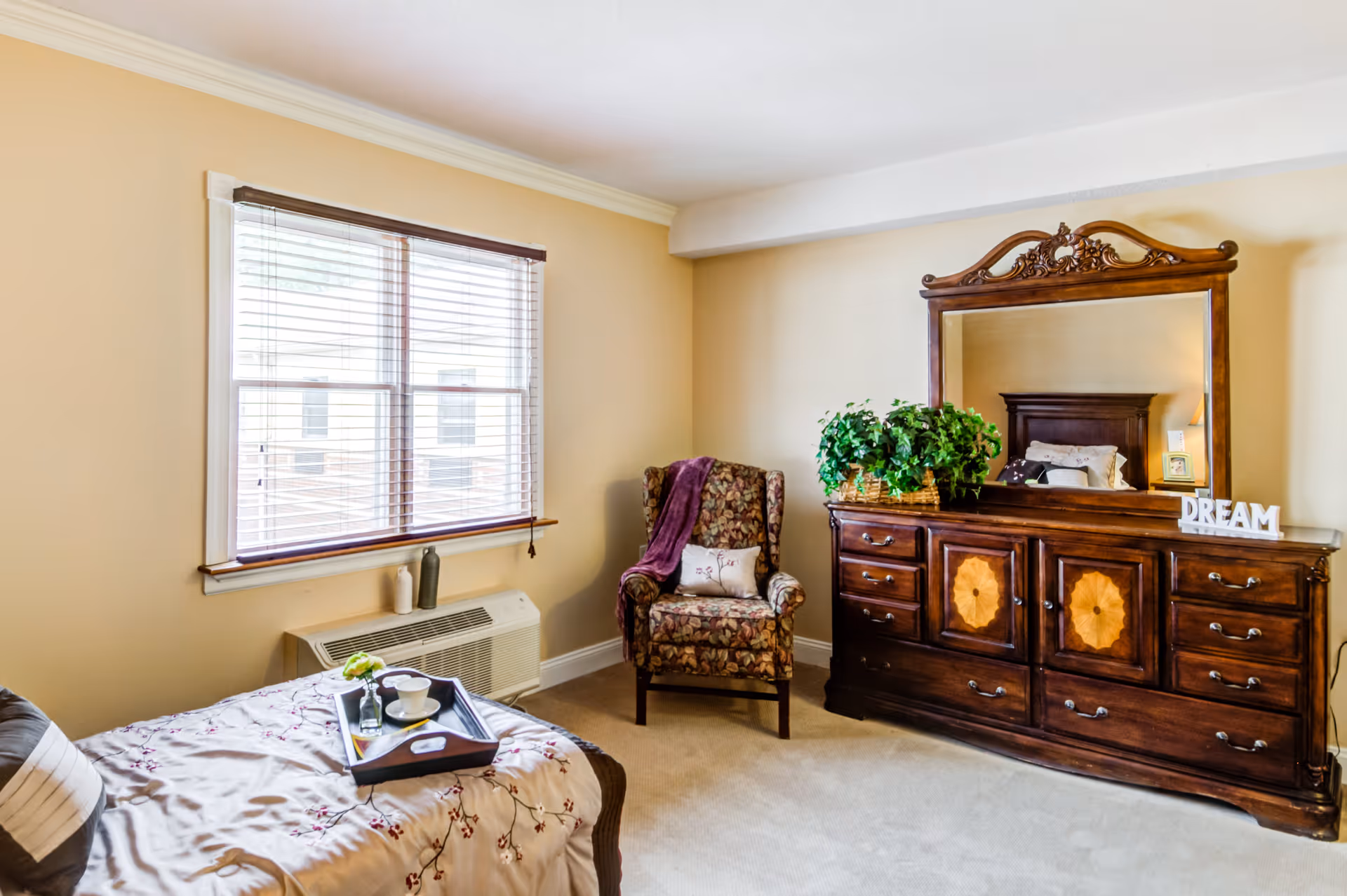 A cozy bedroom with a floral armchair draped with a purple blanket, a wooden dresser with an ornate mirror, a green potted plant, and a decorative 'DREAM' sign. A bed with a floral bedspread and a tray with cups is partially visible. The room has beige walls and a window with blinds letting in natural light.