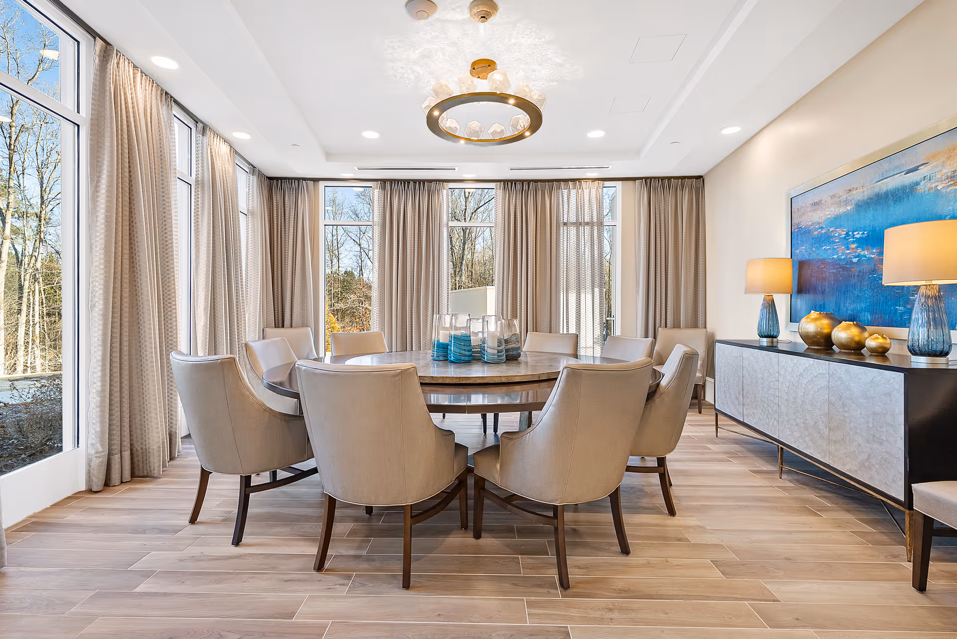 A bright dining room with a large round wooden table surrounded by eight beige upholstered chairs. The room features large windows with beige curtains, allowing natural light to fill the space. A modern circular chandelier hangs above the table. On the right side, there is a sideboard with two blue lamps and decorative golden objects, along with a large blue and gold abstract painting on the wall.
