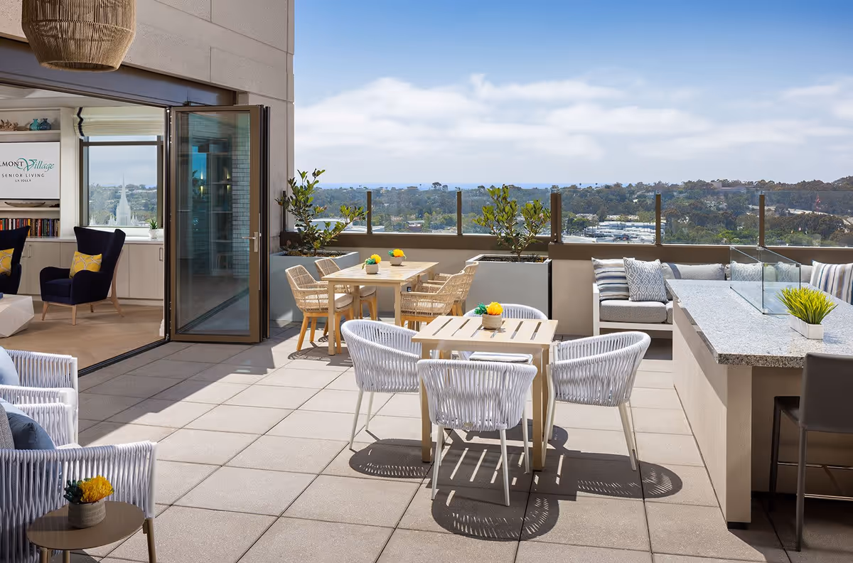 Outdoor patio area at Belmont Village Senior Living La Jolla featuring multiple seating arrangements including white woven chairs around wooden tables, a cushioned bench with pillows, and a granite countertop bar with stools. The patio overlooks a scenic view of trees and distant buildings under a partly cloudy sky. An open glass door leads to an indoor lounge area with navy blue chairs and yellow pillows.