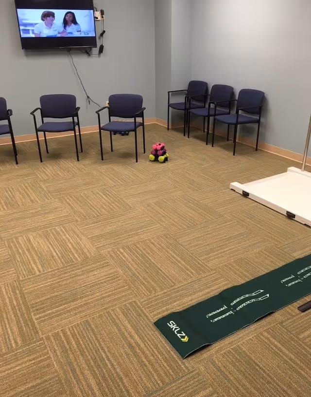 A room with beige patterned carpet, six blue chairs arranged along the walls, a television mounted on the wall showing two people, a small pyramid of colorful balls on the floor, and a green exercise mat labeled 'SKLZ' on the floor.