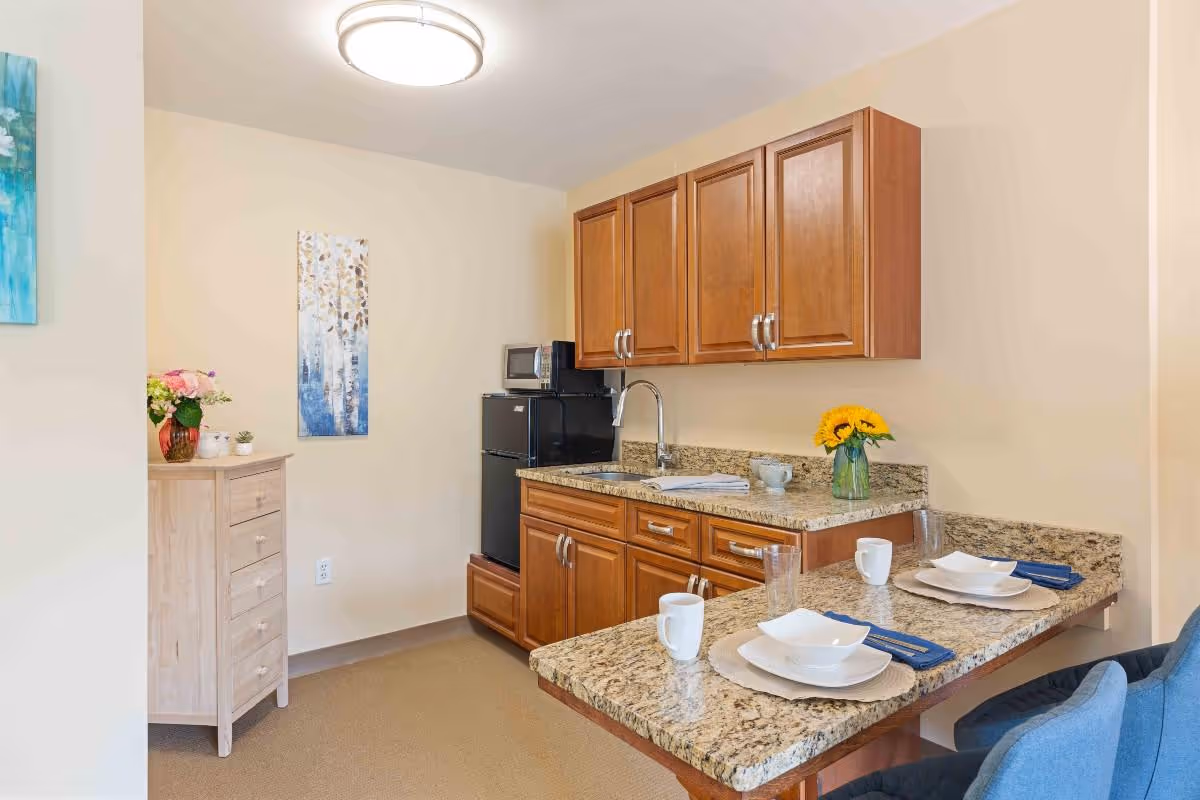 A small kitchen area with wooden cabinets, a granite countertop, a sink, a black mini refrigerator with a microwave on top, and a breakfast bar set with two place settings including white plates, bowls, mugs, and blue napkins. There is a vase with sunflowers on the counter and a small wooden dresser with flowers and small plants on top. The walls are painted beige and there is a piece of artwork hanging on the wall.