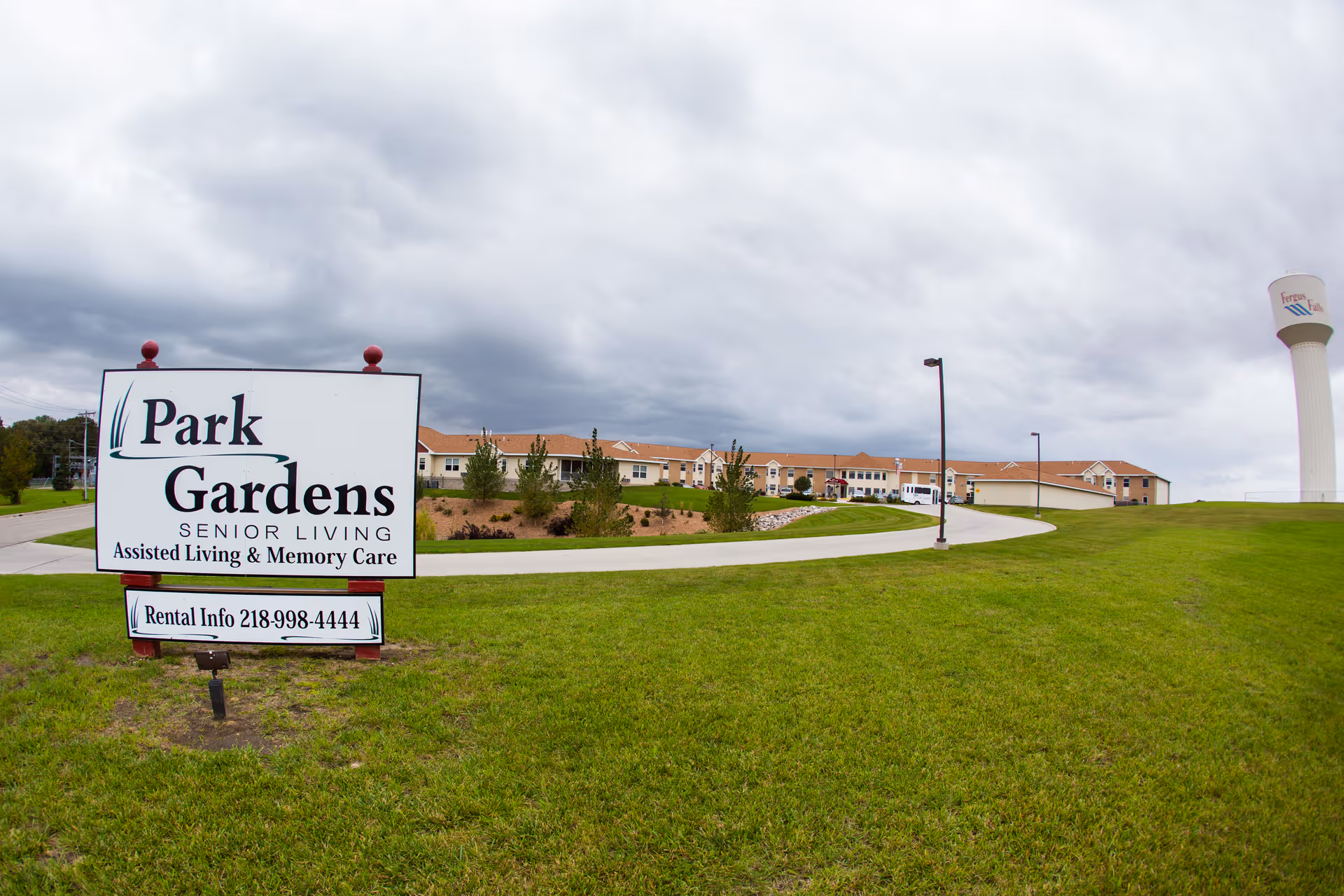 Wide view of the exterior of Park Gardens Senior Living facility with a large sign in the foreground displaying the name, services offered, and rental info phone number. The building is a single-story structure with a tan exterior and red roof, surrounded by green grass and a curved driveway. A water tower is visible in the background under a cloudy sky.