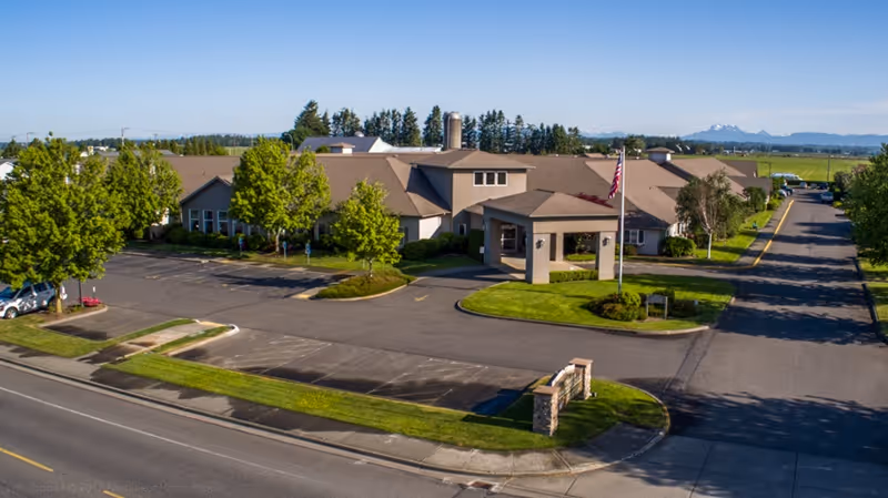 A single-story care facility with a covered front entrance, American flag, parking lot and landscaped grounds under a clear blue sky.