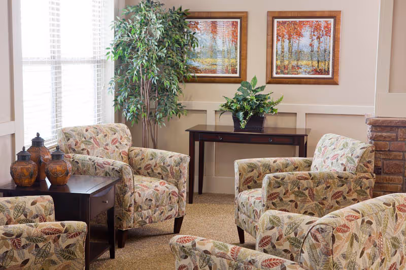 A cozy sitting area with four upholstered armchairs featuring a leaf pattern, arranged around a small dark wooden side table with three decorative urns. Behind the chairs is a dark wooden console table with a green potted plant, flanked by two framed autumn-themed paintings on a beige wall. A tall leafy plant stands near a window with white blinds, and part of a stone fireplace is visible on the right.