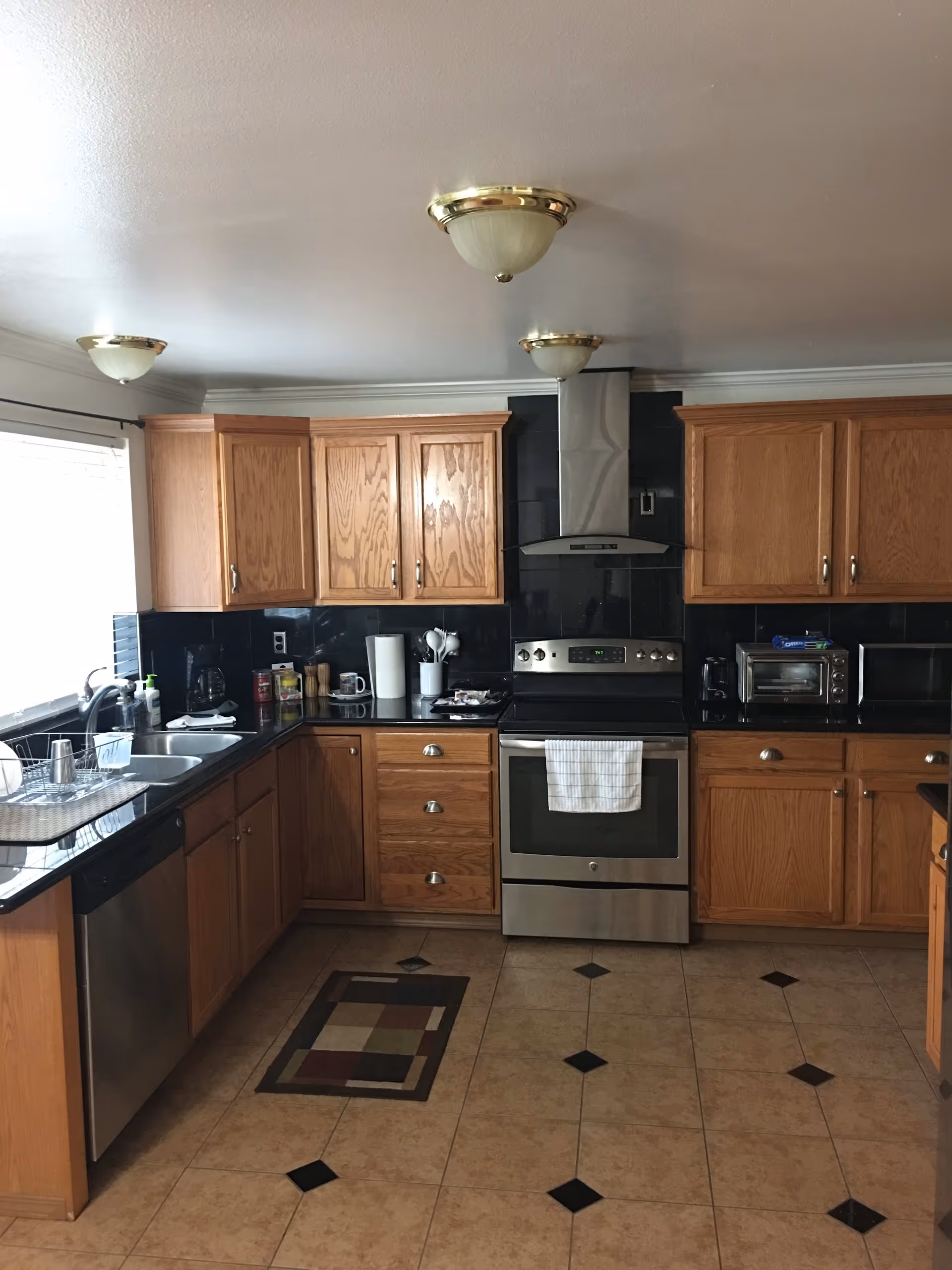 Kitchen with oak cabinets, stainless steel stove and dishwasher, black countertops, and tiled floor with a small rug.