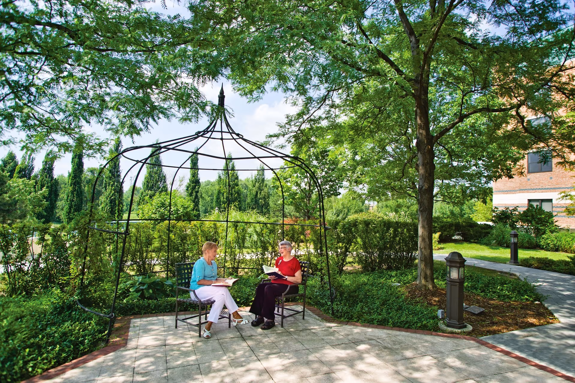 Two elderly women sitting and reading books under a black metal gazebo structure in a lush green garden area with trees, bushes, and a paved walkway nearby. A brick building is partially visible in the background.
