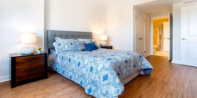A bright bedroom with a queen-sized bed featuring a blue and white patterned bedspread and multiple pillows. There are two dark wooden nightstands on either side of the bed, each with a white lamp. The room has wooden flooring and white walls. In the background, an open door reveals a bathroom with a shower curtain.