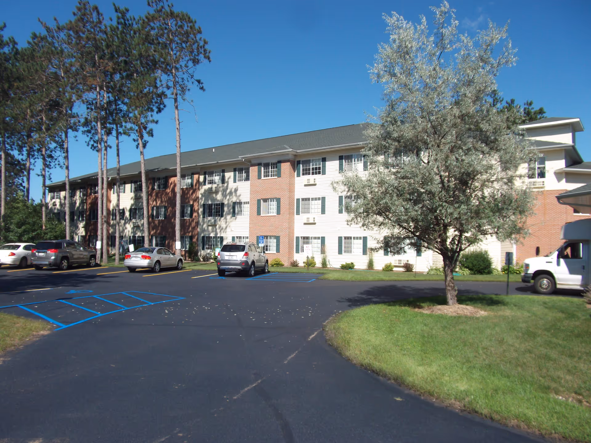 Exterior view of a three-story senior living facility building with a brick and white siding facade, surrounded by trees and a parking lot with several parked cars under a clear blue sky.