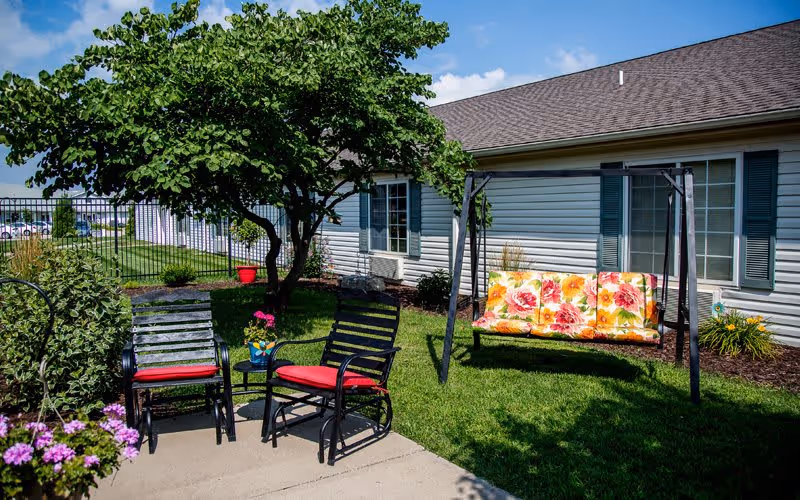 Outdoor patio area with two black metal chairs with red cushions, a small round table with a flower pot, a swing with floral cushions, a tree providing shade, and a building with white siding and green shutters in the background under a blue sky with some clouds.