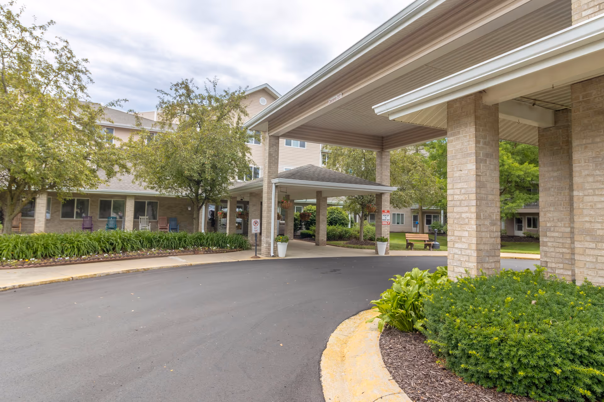 Entrance area of a senior living facility with a covered drop-off zone supported by brick columns. Surrounding the driveway are green shrubs, trees, and a bench. The building has multiple windows and a few colorful rocking chairs on the porch.