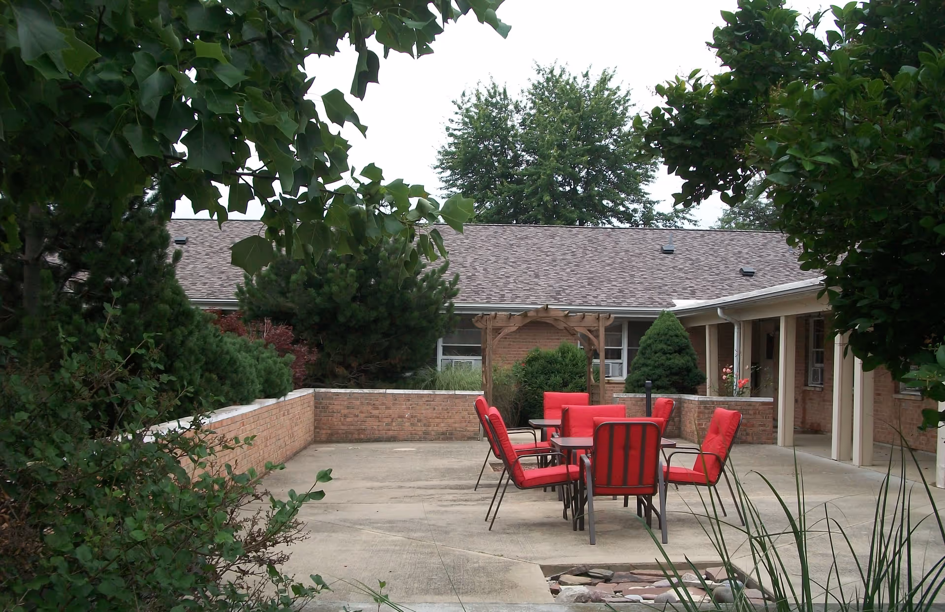 Outdoor patio area at Haven of Arcola with a set of red cushioned chairs around a table on a concrete surface, surrounded by brick walls and greenery including trees and bushes.