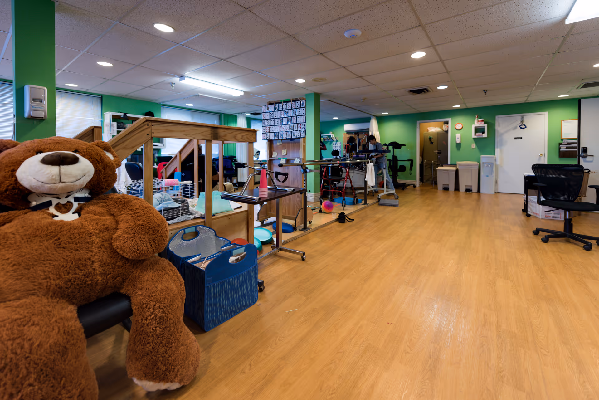 Interior view of a rehabilitation and healing facility room with green walls and wooden flooring. A large brown teddy bear is seated on a chair in the foreground. The room contains various physical therapy equipment, including parallel bars, exercise balls, and walkers. There are chairs, a desk, and a person working in the background near the equipment. The ceiling has recessed lighting and there are windows along the left wall.
