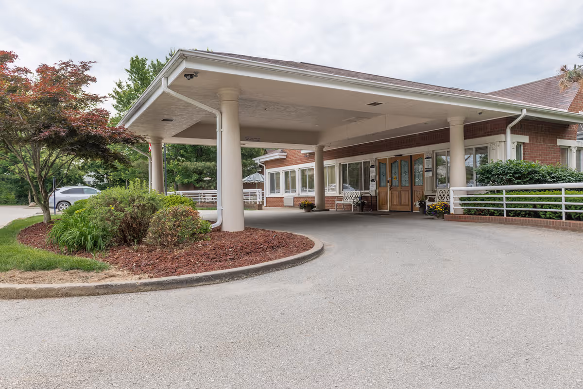 Covered drive-up entrance with columns, double doors, benches and landscaping at the front of a senior living facility.