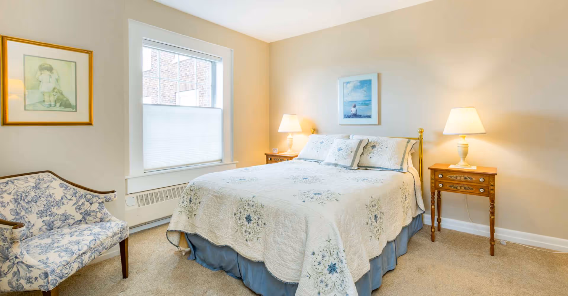 Well-lit bedroom with a double bed dressed in a floral quilt, two nightstands with lamps, a patterned armchair, and a window.