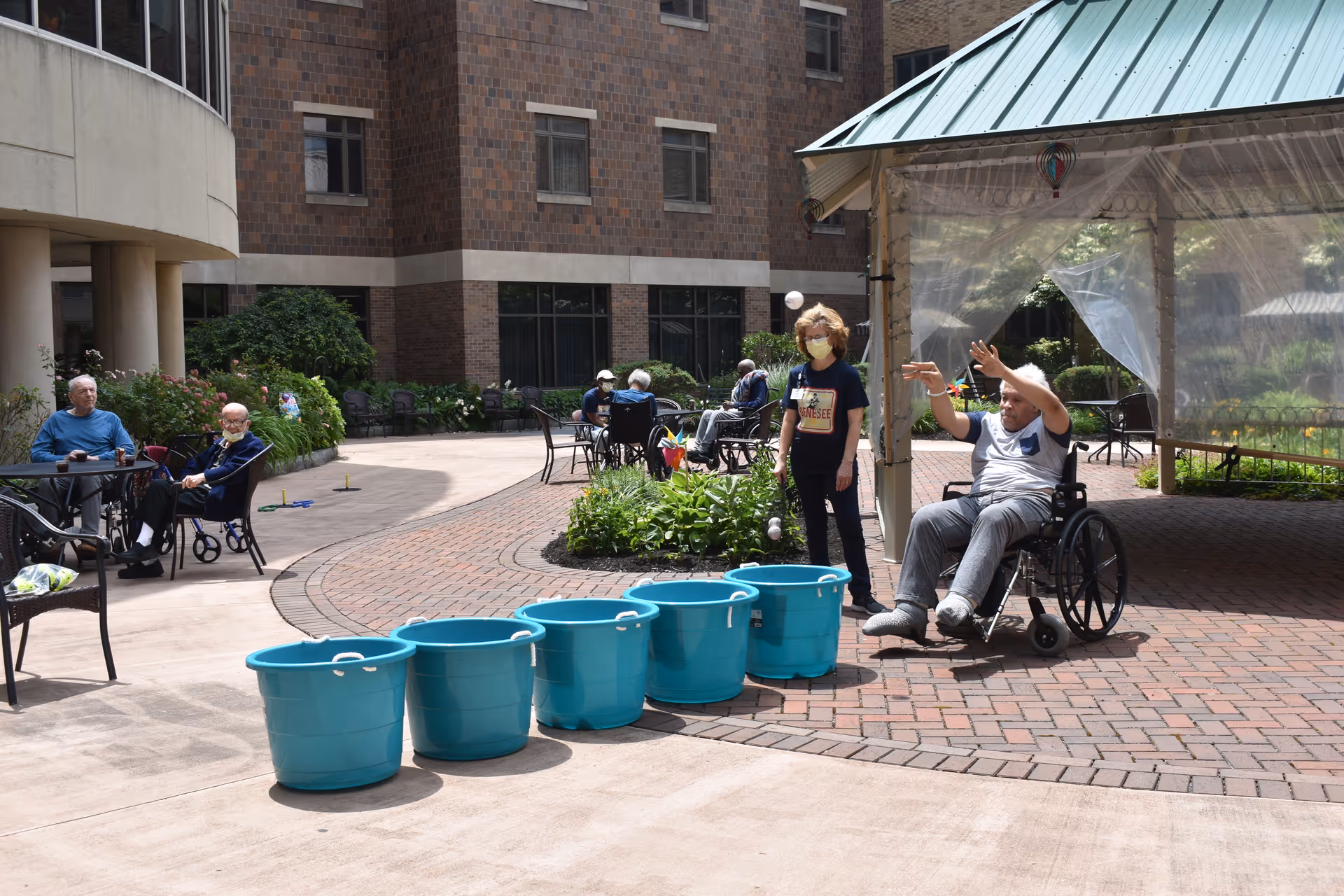 An outdoor courtyard at a senior living facility with several elderly people sitting and socializing. One man in a wheelchair is throwing a ball towards a row of blue buckets, while a staff member wearing a mask stands nearby. The courtyard is surrounded by a multi-story brick building and has paved walkways and garden areas.