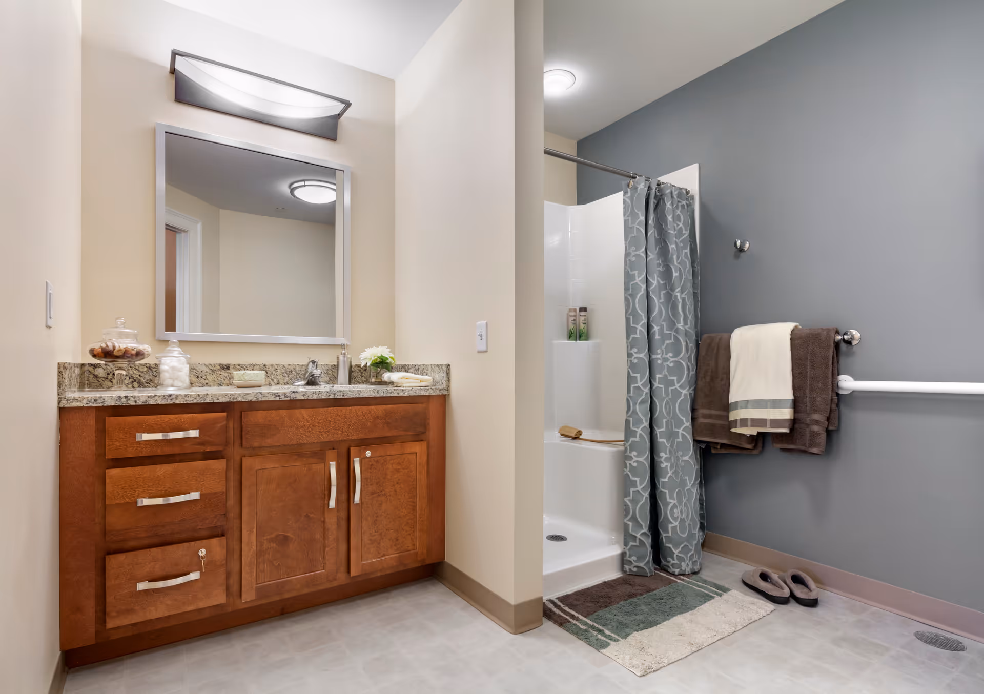 A bathroom featuring a wooden vanity with granite countertop, a large mirror with a light fixture above it, a shower area with a patterned gray shower curtain, towels hanging on a wall-mounted bar, a bath mat, and a pair of slippers on the floor. The walls are painted beige and gray.