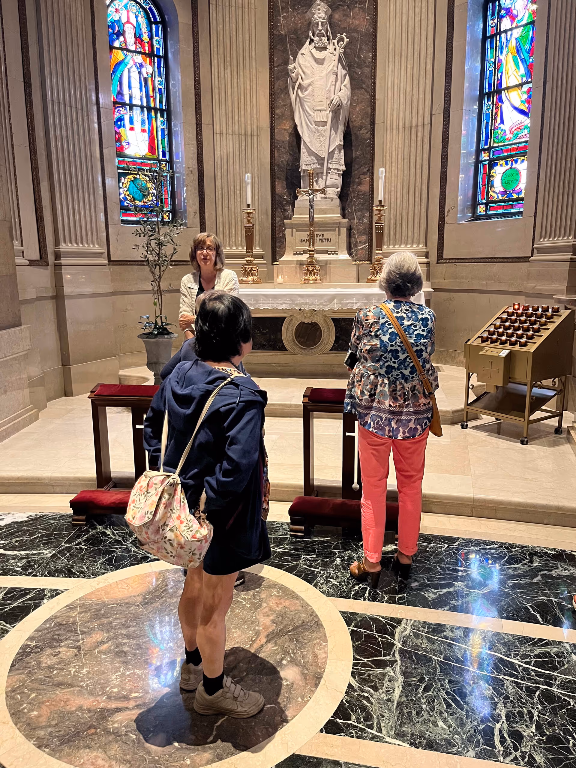 Three women standing inside a chapel or church area with marble floors and stained glass windows. They are facing an altar with a statue of a religious figure, two lit candles, and a crucifix. One woman is speaking while the other two listen.