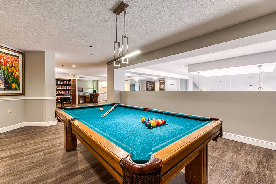 Interior view of a recreational room featuring a pool table with billiard balls and cues set up for a game. The room has wood flooring, neutral-colored walls, and modern lighting fixtures. In the background, there is a bookshelf filled with books and a desk with a computer, suggesting a multi-purpose common area.