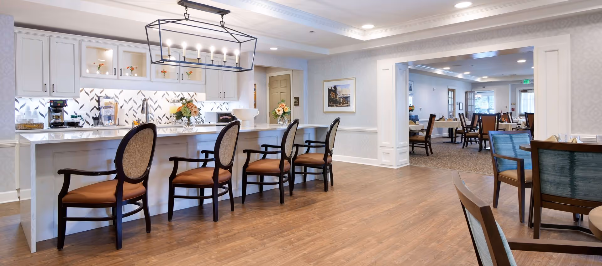 Open, well-lit communal dining area with a white kitchen island, cushioned wooden bar stools, and tables in an adjacent dining room.