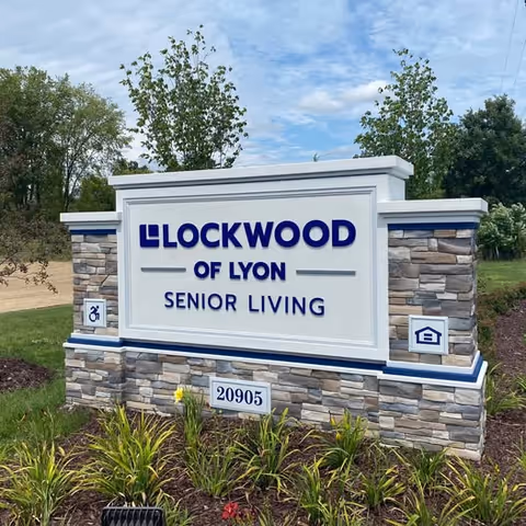 Outdoor stone and white sign for Lockwood of Lyon Senior Living with the address number 20905, surrounded by green plants and trees under a partly cloudy sky.