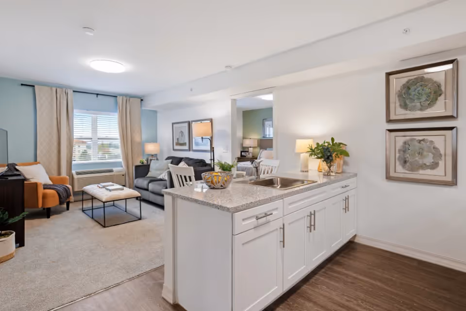 Open-plan living area with a kitchen island in the foreground, a sofa and armchair near a large window, and framed artwork on the wall.
