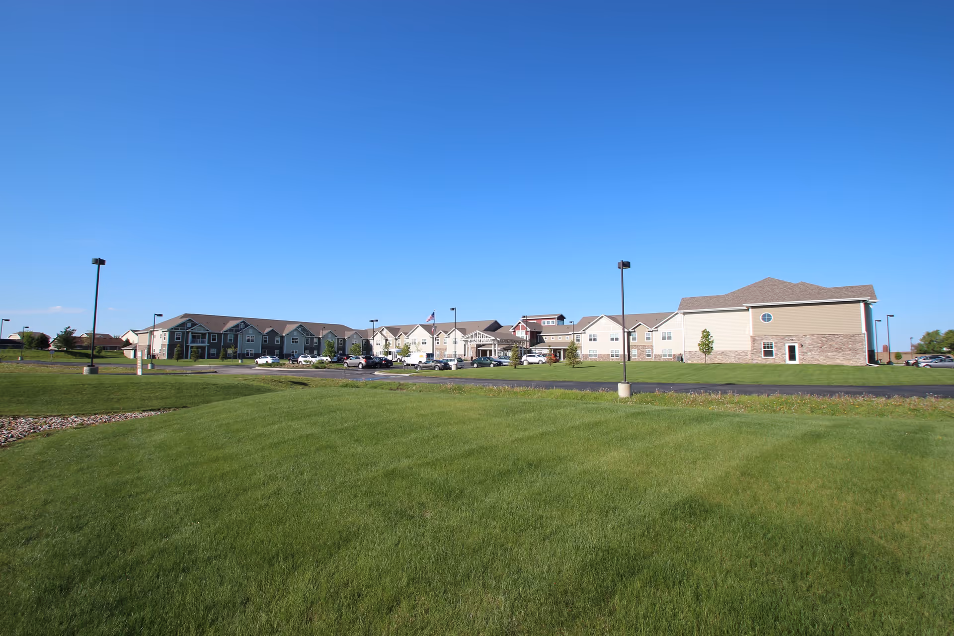 Expansive green lawn leading to a low-rise senior living complex and parking area under a clear blue sky.