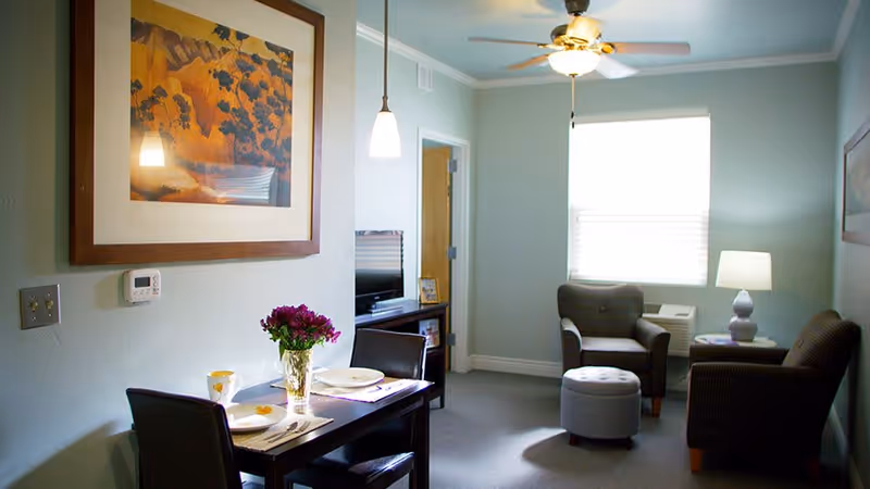 A cozy interior room with a small dining table set for two, featuring plates, cups, and a vase with purple flowers. The room also includes two armchairs, a small ottoman, a side table with a lamp, a wall-mounted thermostat, a ceiling fan with a light, and a window with blinds letting in natural light. A framed artwork hangs on the wall above the dining table.